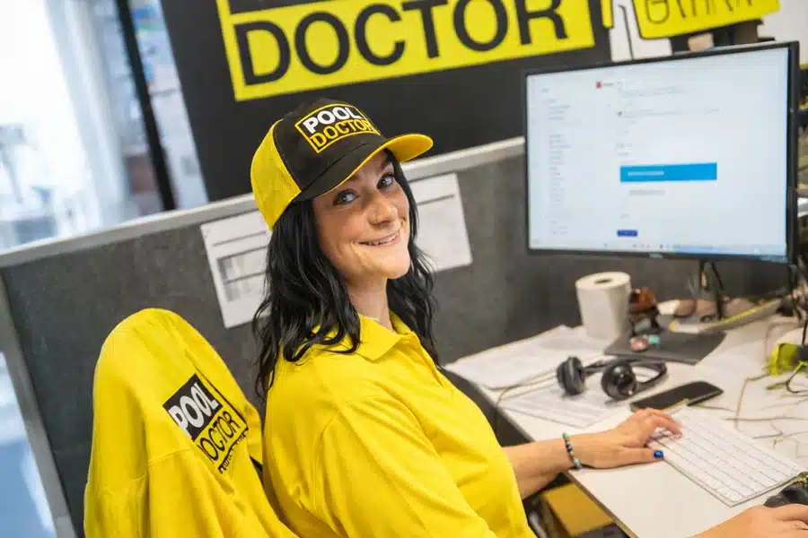 A woman wearing a yellow "Pool Doctor" uniform and cap sits at an office desk, smiling at the camera while working on a computer. Headset, keyboard, and pool maintenance paperwork are on the desk. Bold "DOCTOR" sign is visible behind her.