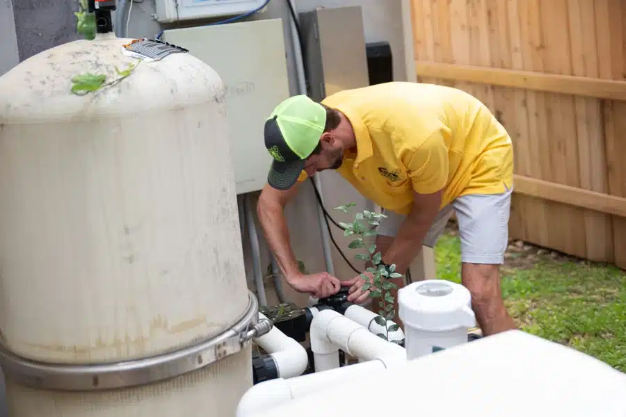 A man in a yellow shirt and green cap is providing equipment care as he inspects pool equipment in a fenced backyard area, leaning over pipes and a large filter tank with tools in hand.
