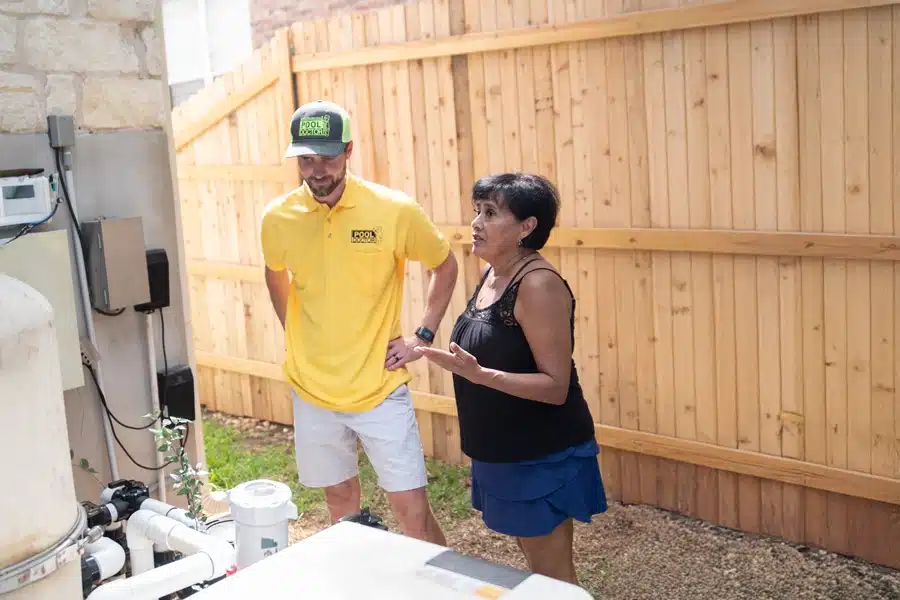 A man in a yellow polo shirt and cap stands next to a woman in a black tank top and blue skirt in a backyard, looking at pool equipment near a wooden fence—true technicians you can trust.